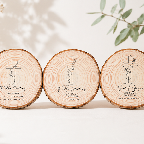 Three round natural wood slice coasters or trivets with engraved Christian crosses and personalized text, arranged side by side on a light neutral surface with soft leaf shadows and eucalyptus branches in the background.