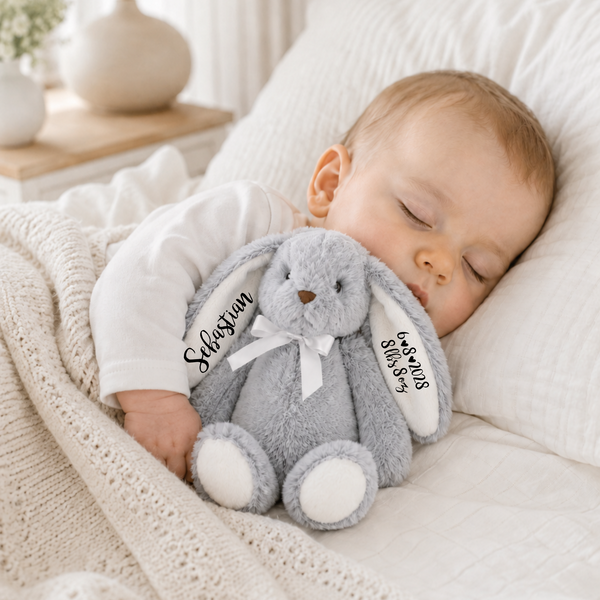 A sleeping baby in a white onesie rests on a white pillow and cream blanket, holding a gray plush bunny toy with white embroidered names on its ears.