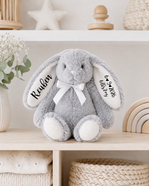 A plush gray bunny toy with long floppy ears sits on a light wood shelf. The left ear is embroidered with the name 'Reuben' and the right ear has a date, and the bunny wears a white ribbon bow.