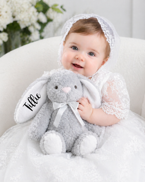 A baby wearing a white lace bonnet and dress sits on a cream chair holding a gray plush bunny stuffed animal with the name 'Tillie' embroidered on the ear.