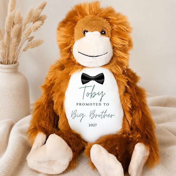 A plush monkey toy with brown fur, a white face and belly, and a black bow tie, sitting on a cream fabric surface. The belly is printed with the text 'Toby PROMOTED TO Big Brother 2027' in teal and black.