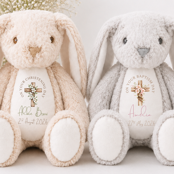 Two plush Easter bunny toys side by side, one cream and one light gray, each with a custom printed white belly area featuring a cross, floral design, and personalized baptism or christening text.