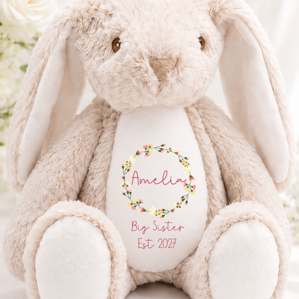 A plush beige bunny toy with long floppy ears sits centered on white fabric, surrounded by white roses. The bunny has a white belly patch with a floral wreath and the text 'Amelie's Big Sister Est 2027' printed on it. The can be personalised any text you'd like
