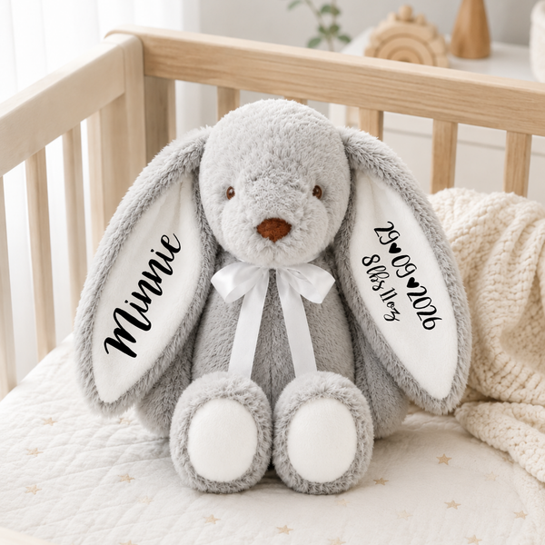 A personalized plush gray bunny stuffed animal sitting in a wooden crib. The bunny has long floppy ears with custom black text on the inside, reading 'Minnie' on the left ear and a date on the right.