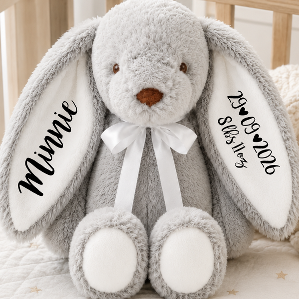 A personalized plush gray bunny stuffed animal sitting in a wooden crib. The bunny has long floppy ears with custom black text on white inner ears reading 'Minnie' and a date, a white satin bow around its neck, and white paw pads.